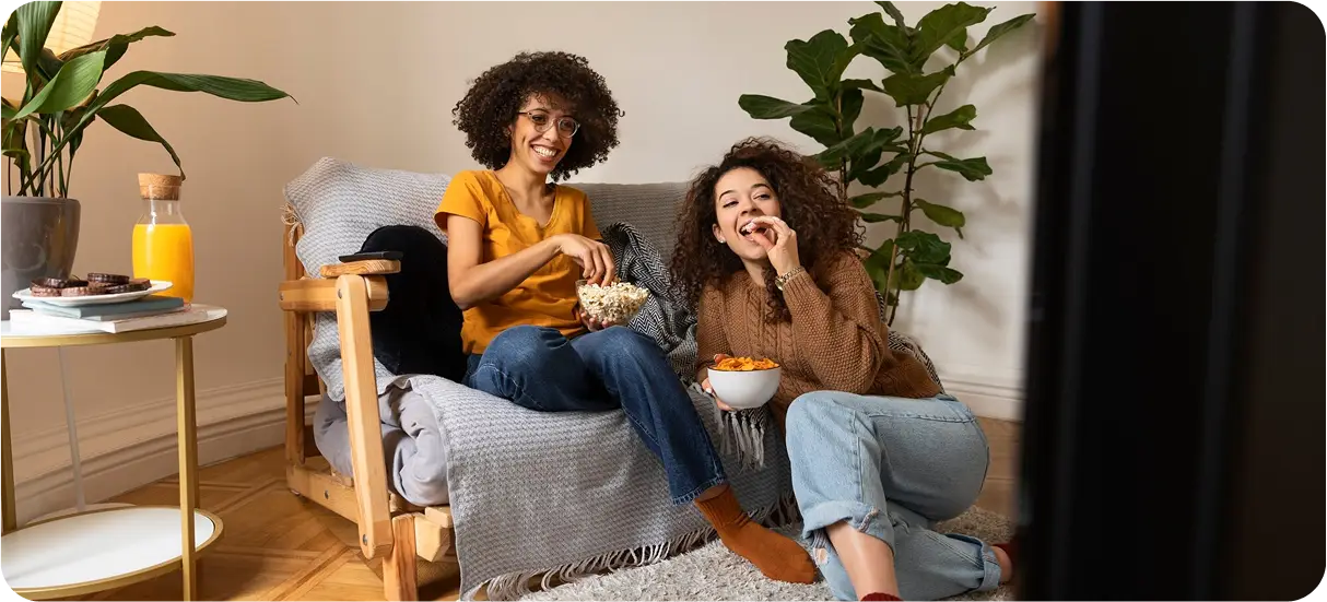 Two friends laughing on a sofa with a bowl of popcorn